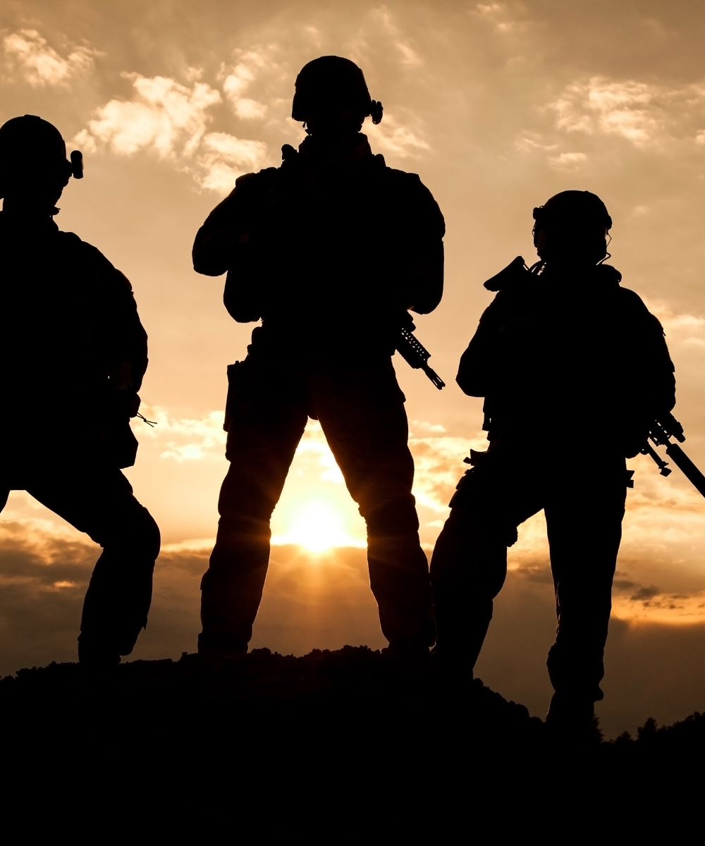 Drei Soldaten stehen silhouettenartig auf einem Hügel im Sonnenuntergang – symbolträchtige Szene für Kameradschaft, Einsatz und Militärgeschichte